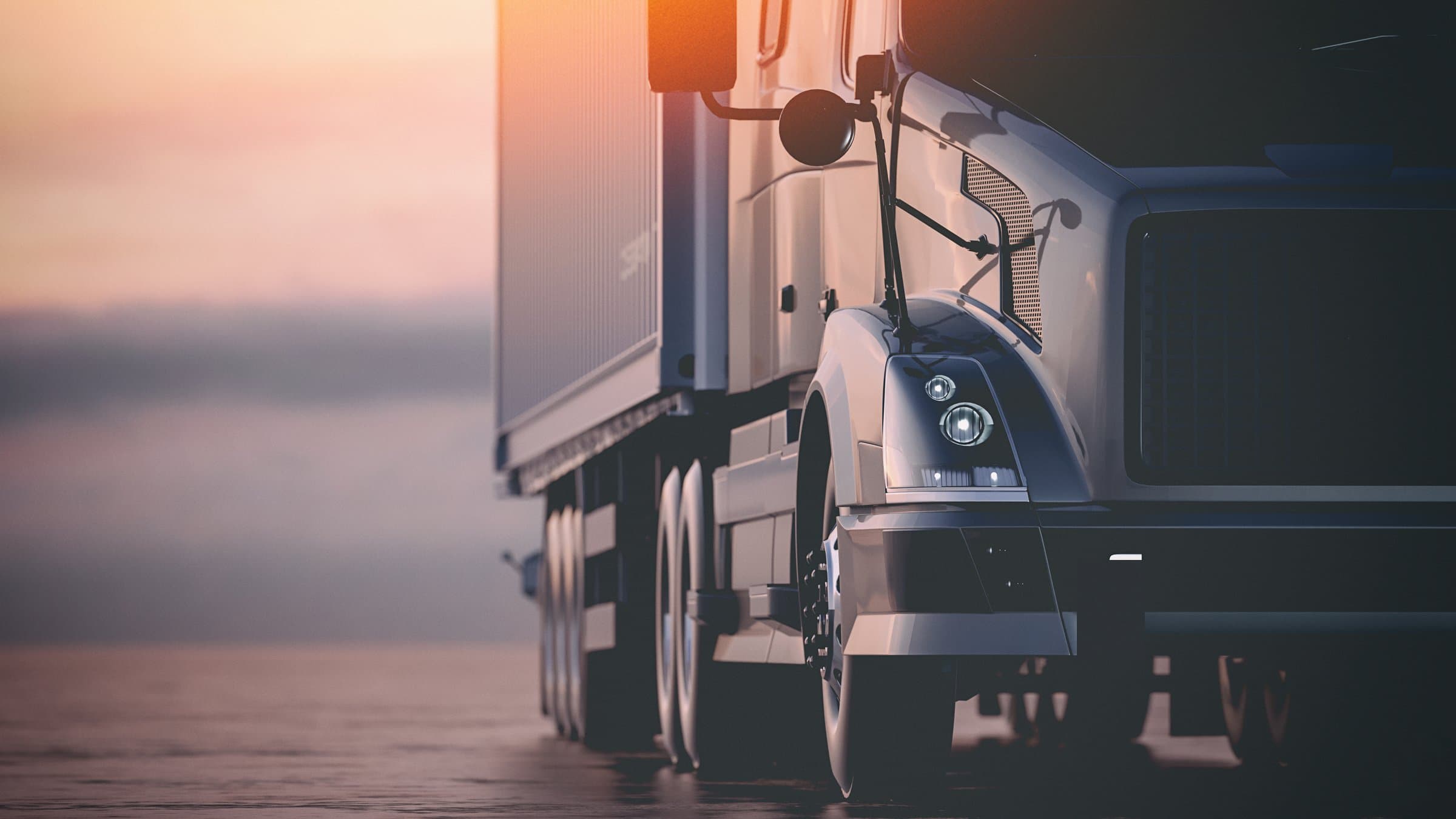 The dark front of a semi truck at dusk, headlight, mirror and grille, with the horizon behind it